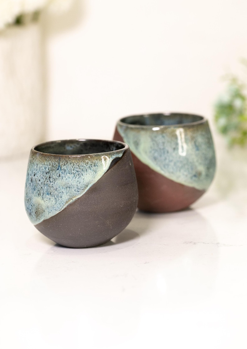 Pu&ograve; includere: Due bicchieri da vino senza stelo in ceramica fatti a mano. La met&agrave; inferiore di ogni bicchiere &egrave; marrone scuro, mentre la met&agrave; superiore &egrave; azzurra e verde. I bicchieri sono su una superficie bianca.