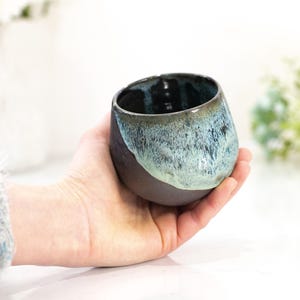 Pu&ograve; includere: Una tazza in ceramica fatta a mano con un design unico. La tazza presenta una base marrone scuro che sfuma in una glassa turchese e blu nella met&agrave; superiore. La tazza &egrave; tenuta in mano, mostrando le sue dimensioni e la sua forma.