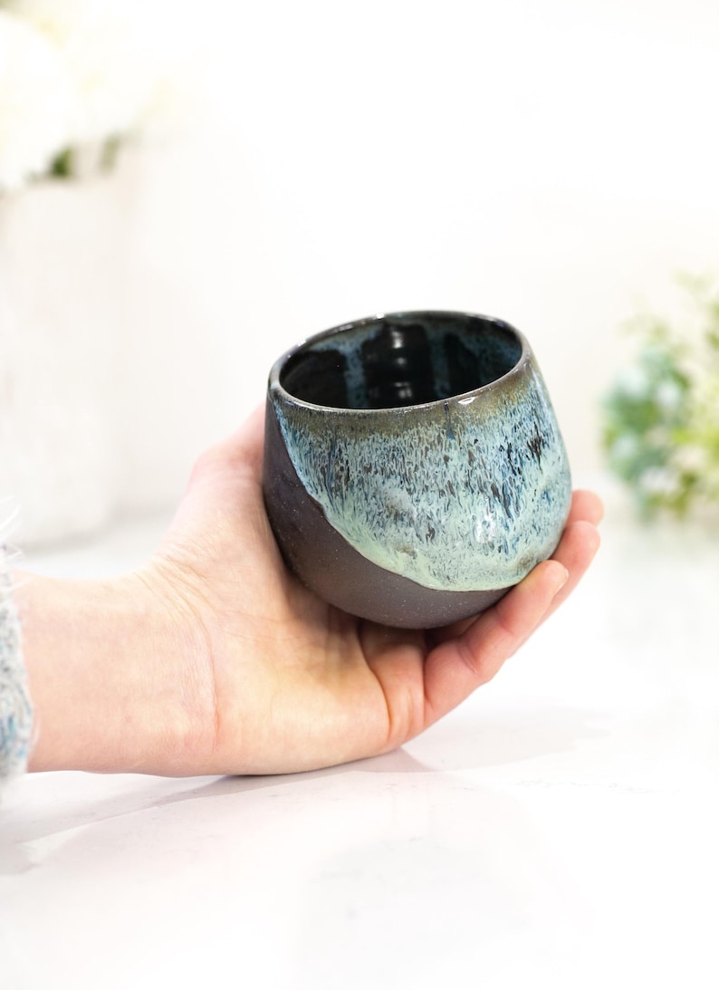 Pu&ograve; includere: Una tazza in ceramica fatta a mano con un design unico. La tazza presenta una base marrone scuro che sfuma in una glassa turchese e blu nella met&agrave; superiore. La tazza &egrave; tenuta in mano, mostrando le sue dimensioni e la sua forma.