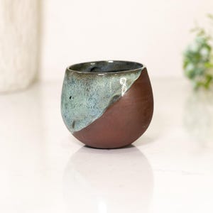Pu&ograve; includere: Una tazza in ceramica fatta a mano con un design unico. La tazza ha una forma arrotondata, con una divisione diagonale di colore. Un lato &egrave; marrone scuro, l'altro &egrave; un misto di verde acqua e blu. La tazza viene probabilmente utilizzata per bere.