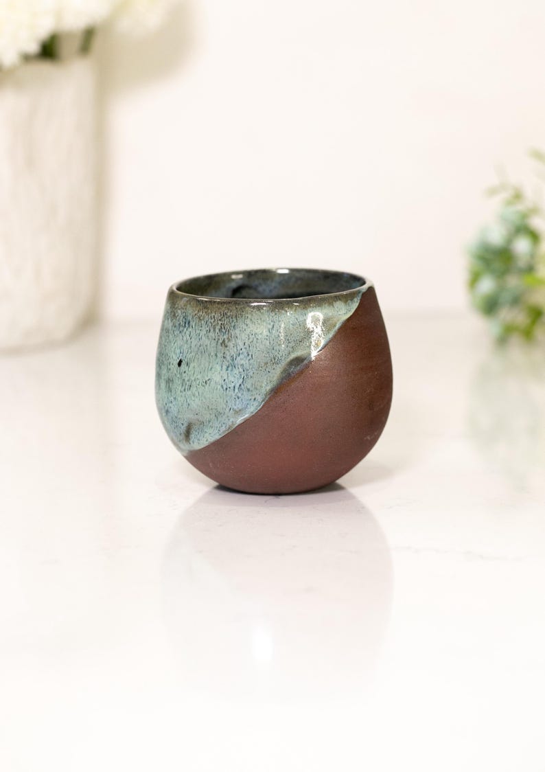 Pu&ograve; includere: Una tazza in ceramica fatta a mano con un design unico. La tazza ha una forma arrotondata, con una divisione diagonale di colore. Un lato &egrave; marrone scuro, l'altro &egrave; un misto di verde acqua e blu. La tazza viene probabilmente utilizzata per bere.