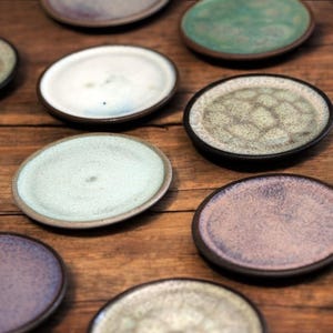 Könnte beinhalten: Eine Sammlung kleiner, runder Keramikplatten in verschiedenen Farben, darunter Weiß, Grün und Lila, mit dunkelbraunen Rändern. Die Teller sind auf einer Holzoberfläche angeordnet und zeigen ihre einzigartigen Texturen und Oberflächen.