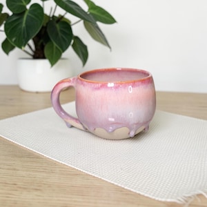 May include: A handcrafted ceramic mug with a pink and purple glaze. The mug features a rounded form, a curved handle, and small feet. The glaze drips down the sides, creating a unique texture. The mug is on a white mat.