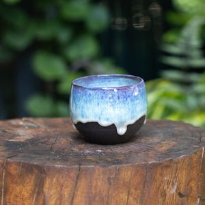 May include: A small, round ceramic cup featuring a matte black base and a glossy, drippy glaze in shades of light blue and purple at the top. The rim is smooth and circular, with the glaze creating a distinct, uneven drip pattern over the dark body.