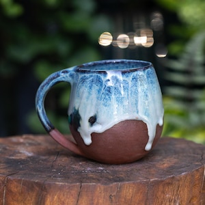 May include: A rounded ceramic mug featuring a dark brown, unglazed base and a glossy, drippy glaze in shades of light blue, white, and deep navy. The mug has a curved handle and a wide, circular opening, showcasing a distinct contrast between the textured clay bottom and the smooth, flowing glaze finish on the upper portion.