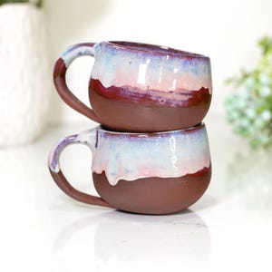 May include: Two handmade ceramic mugs stacked on top of each other. The mugs have a dark brown base with a gradient of colors on the upper half, including shades of red, pink, and blue. Each mug has a rounded handle.