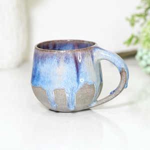 May include: A ceramic mug with a rounded shape and a handle. The mug has a gradient of colours, with a dark brown rim, transitioning to blue and white, and a grey base. The glaze drips down the sides, creating a unique texture.