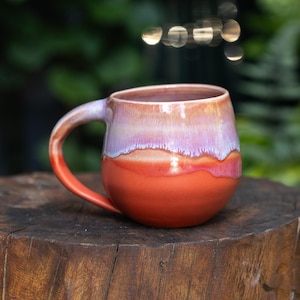 Può includere: Una tazza in ceramica arrotondata con una smaltatura liscia e lucida. La parte superiore è di un delicato color lavanda che sfuma in una base arancione vibrante e solida. La tazza presenta un manico curvo e un bordo ampio e aperto.