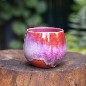 May include: A rounded ceramic cup featuring a vibrant glaze that transitions from a deep red at the top to a bright magenta in the middle, with a raw, unglazed terracotta base visible at the bottom, creating a distinct contrast in texture and colour.