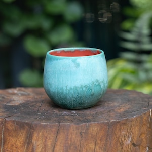May include: A rounded ceramic cup featuring a textured turquoise and teal glaze on the exterior with a smooth, solid orange interior. The vessel has a wide, open top and a curved body that tapers slightly towards the base, showcasing a reactive finish.