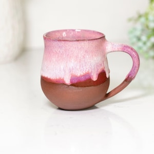 May include: A handmade ceramic mug with a rounded base and a handle. The mug features a gradient of colors, transitioning from a deep brown at the base to a light pink at the rim, with a darker pink band in between. The mug is suitable for hot or cold beverages.