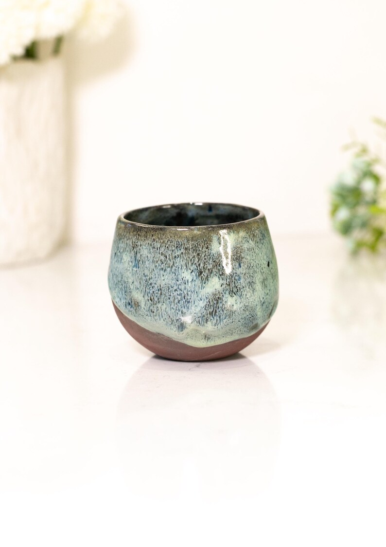 Pu&ograve; includere: Un bicchiere in ceramica fatto a mano con una forma organica unica. L'esterno presenta una miscela di smalto blu e verde su una base marrone scuro. L'interno &egrave; blu scuro. Ideale per le bevande.