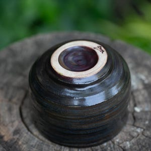 May include: A handmade ceramic bowl, viewed from the bottom, showcasing a dark, glossy exterior with subtle horizontal striations. The interior is a deep burgundy, and the rim is a creamy white. The bowl rests on a weathered wooden surface.