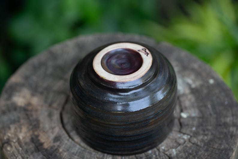 May include: A handmade ceramic bowl, viewed from the bottom, showcasing a dark, glossy exterior with subtle horizontal striations. The interior is a deep burgundy, and the rim is a creamy white. The bowl rests on a weathered wooden surface.