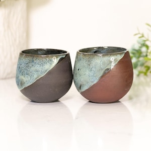 Pu&ograve; includere: Due bicchieri da vino senza stelo in ceramica fatti a mano. Ogni bicchiere presenta un design unico, con una divisione diagonale marrone scuro e verde acqua. I bicchieri sono rotondi e hanno una finitura liscia e lucida.