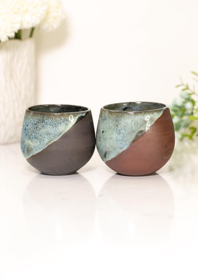 Pu&ograve; includere: Due bicchieri da vino senza stelo in ceramica fatti a mano. Ogni bicchiere presenta un design unico, con una divisione diagonale marrone scuro e verde acqua. I bicchieri sono rotondi e hanno una finitura liscia e lucida.