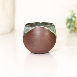 Pu&ograve; includere: Una tazza in ceramica fatta a mano con una forma arrotondata. La parte inferiore &egrave; marrone scuro, con un bordo verde-acqua. La tazza viene probabilmente utilizzata per bere bevande.