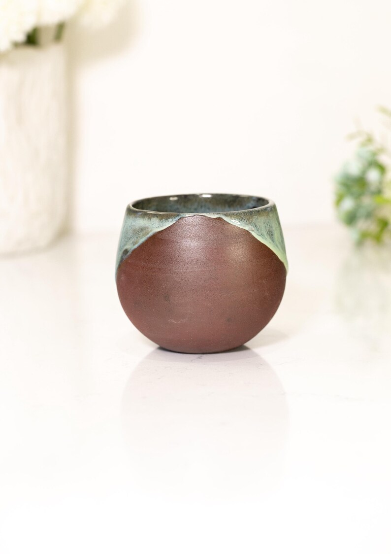 Pu&ograve; includere: Una tazza in ceramica fatta a mano con una forma arrotondata. La parte inferiore &egrave; marrone scuro, con un bordo verde-acqua. La tazza viene probabilmente utilizzata per bere bevande.