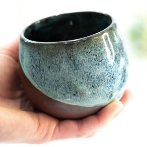 Pu&ograve; includere: Una tazza in ceramica fatta a mano con una forma arrotondata. La met&agrave; superiore presenta una smaltatura blu-verde screziata, che sfuma in una base marrone scuro. La tazza &egrave; tenuta in una mano su sfondo bianco.
