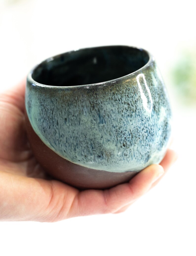 Pu&ograve; includere: Una tazza in ceramica fatta a mano con una forma arrotondata. La met&agrave; superiore presenta una smaltatura blu-verde screziata, che sfuma in una base marrone scuro. La tazza &egrave; tenuta in una mano su sfondo bianco.
