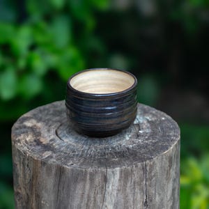 May include: A small, handcrafted ceramic bowl with a dark blue and brown exterior and a light beige interior. The bowl is cylindrical with a slightly flared rim. It sits on a weathered wooden stump against a blurred green background.