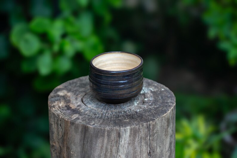 May include: A small, handcrafted ceramic bowl with a dark blue and brown exterior and a light beige interior. The bowl is cylindrical with a slightly flared rim. It sits on a weathered wooden stump against a blurred green background.