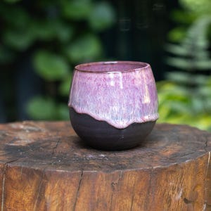 Pode incluir: Uma chávena de cerâmica arredondada com uma base preta mate e um acabamento de esmalte brilhante e escorrido em tons de rosa vibrante e lavanda suave. A parte superior do recipiente apresenta uma transição fluida onde o esmalte rosa sobrepõe a argila.