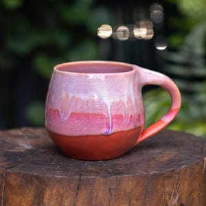 Peut inclure: Une tasse en céramique arrondie dotée d'un émail brillant passant d'un rose pâle au bord à un magenta vif au milieu, se terminant par une base en terre cuite mate unie. La tasse comprend une anse incurvée et une forme de corps lisse et bombée.