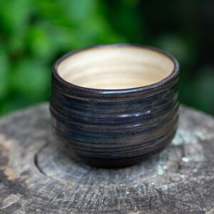 May include: A handmade ceramic bowl with a dark blue and brown exterior and a light beige interior. The bowl has a cylindrical shape with horizontal ridges and sits on a wooden surface. The background is a blurred green.