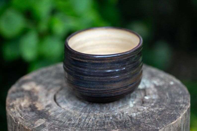 May include: A handmade ceramic bowl with a dark blue and brown exterior and a light beige interior. The bowl has a cylindrical shape with horizontal ridges and sits on a wooden surface. The background is a blurred green.