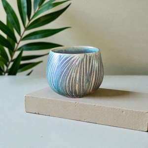 Peut inclure: Une tasse en céramique artisanale avec un design texturé unique. La tasse présente un mélange de couleurs bleu, turquoise et or, avec des lignes verticales créant un motif rayé. La tasse est posée sur un bloc rectangulaire gris, avec une plante verte feuillue en arrière-plan.