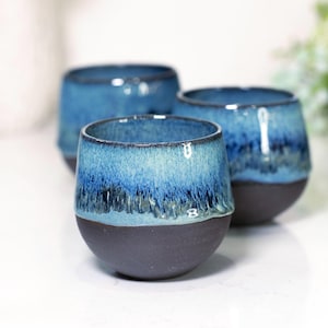 Op de afbeelding: Drie handgemaakte keramische bekers met een tweekleurig glazuur. De bovenste helft van elke beker is een levendig blauw, dat overgaat in een donkerder blauw aan de rand. De onderste helft is donkergrijs. Ideaal voor koffie of thee.