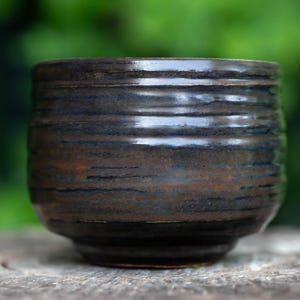May include: A handmade ceramic cup with a dark brown and blue glaze. The cup has a rounded shape with horizontal textured lines. The cup is sitting on a wooden surface, with a blurred green background.