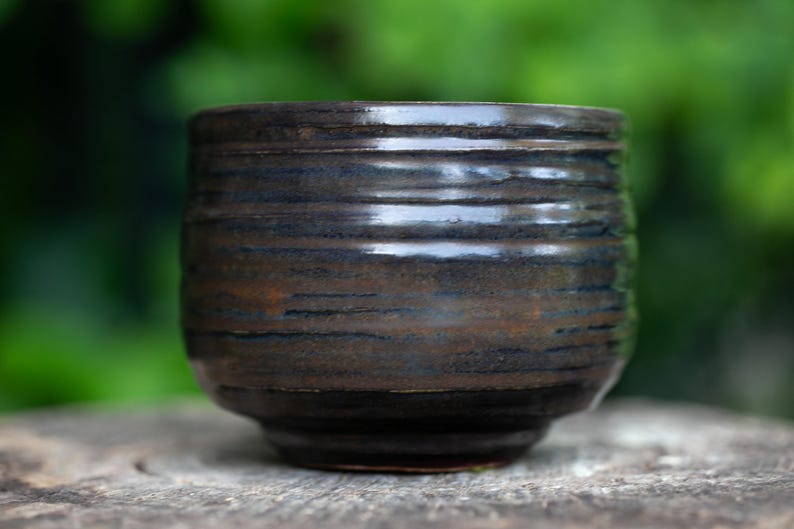 May include: A handmade ceramic cup with a dark brown and blue glaze. The cup has a rounded shape with horizontal textured lines. The cup is sitting on a wooden surface, with a blurred green background.