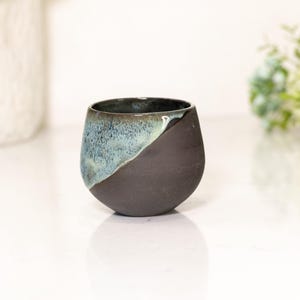 Pu&ograve; includere: Una tazza in ceramica fatta a mano con un design unico. La tazza &egrave; di colore marrone scuro nella met&agrave; inferiore e passa a una glassa blu-verde nella met&agrave; superiore. Ha una forma arrotondata e una finitura liscia.