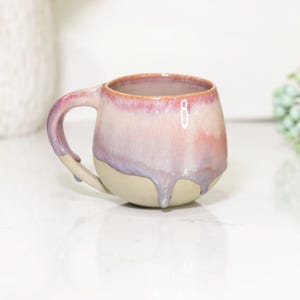 Puede incluir: Taza de cerámica hecha a mano con forma redondeada y asa. La taza presenta un degradado de colores, incluyendo rosa, morado y beige. El esmalte tiene un efecto goteado, lo que añade a su apariencia única.