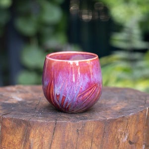 May include: A rounded ceramic cup featuring a glossy glaze with blended shades of vibrant magenta, deep red, and streaks of violet. The surface displays a subtle carved leaf-like pattern that radiates from the base towards the rim of the vessel.