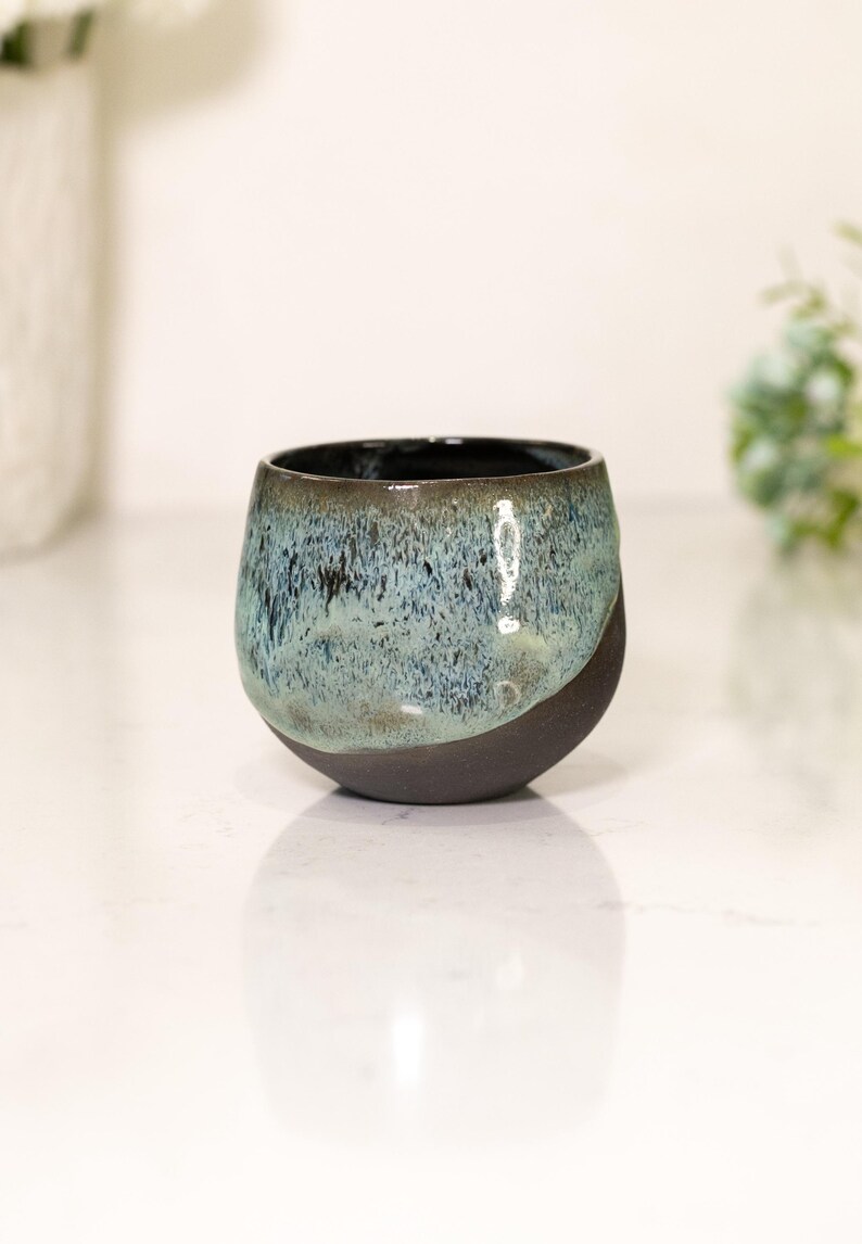 Pu&ograve; includere: Una ciotola in ceramica fatta a mano con un design unico. La ciotola presenta una base nera opaca scura che si trasforma in una glassa screziata blu e turchese. L'interno &egrave; di colore scuro e lucido.