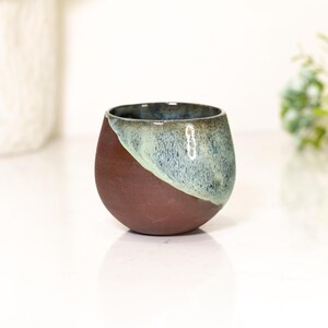 Pu&ograve; includere: Una piccola tazza rotonda in ceramica con una combinazione di colori unica. La met&agrave; inferiore &egrave; di un marrone rossastro scuro, mentre la met&agrave; superiore &egrave; una miscela diagonale di blu e verde. La tazza ha una finitura liscia e lucida.