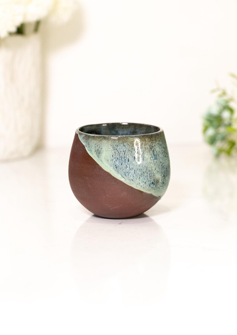Pu&ograve; includere: Una piccola tazza rotonda in ceramica con una combinazione di colori unica. La met&agrave; inferiore &egrave; di un marrone rossastro scuro, mentre la met&agrave; superiore &egrave; una miscela diagonale di blu e verde. La tazza ha una finitura liscia e lucida.