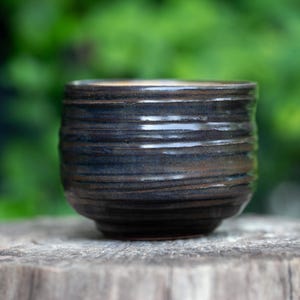 May include: A handmade, dark brown ceramic cup with a textured, horizontal ridged design. The cup is cylindrical with a slightly rounded base. The background is a blurred green, suggesting an outdoor setting. The cup is sitting on a wooden surface.