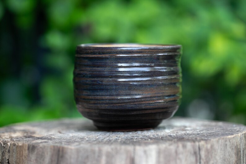 May include: A handmade, dark brown ceramic cup with a textured, horizontal ridged design. The cup is cylindrical with a slightly rounded base. The background is a blurred green, suggesting an outdoor setting. The cup is sitting on a wooden surface.