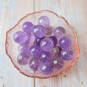 May include: A collection of polished amethyst spheres displayed in a pink, scalloped-edge glass bowl with a gold rim. The spheres vary in shades of purple and are arranged on a light wood-grain surface.