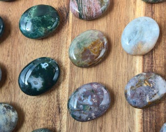 May include: A selection of polished gemstones in shades of green, brown, and pink, arranged on a wooden surface. The stones are smooth, oval-shaped, and feature natural patterns, ideal for jewellery or decorative purposes.