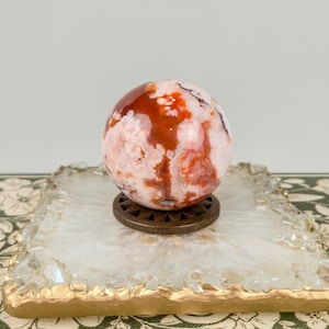 May include: A polished spherical stone featuring a marbled pattern of deep red, orange, and creamy white inclusions. The sphere rests on a small, dark brown circular stand with a sunburst-like carved design, placed atop a square white geode coaster.