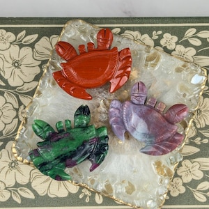May include: Three carved stone crab figurines arranged on a square, translucent, textured tray with gold-toned edges. One crab is solid red, one is deep green with dark inclusions, and the third is a variegated blend of purple, grey, and red tones.