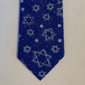 May include: A blue necktie featuring a pattern of white Star of David symbols and small white dots. The tie is made of a smooth fabric and is displayed against a white background. The tie's design is a repeating pattern of the Star of David.