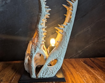 May include: An off-white alligator skull lamp with prominent teeth and an internal light bulb. The lamp is mounted on a black base, contrasting with the dark backdrop and wooden surface. The light highlights the skull's intricate details.