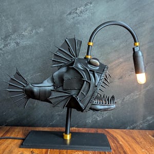 May include: A black anglerfish lamp with a curved arm and a lightbulb. The lamp features a detailed, textured design with fins, teeth, and a base. Gold accents are visible on the lamp's arm and base. The lamp is on a black rectangular base.