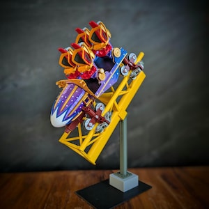 May include: A miniature roller coaster model with a yellow track, a purple and white car, and red and gold accents. The coaster is mounted on a gray and black base, set against a dark background. The model is a detailed representation of a theme park ride.